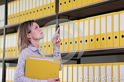 Attentive female clerk counts folders on rack in archive Stock Photo