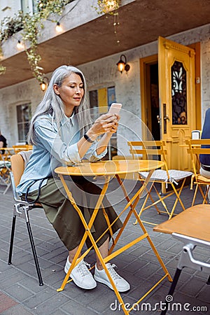 Attracive long haired senior Asian lady holds mobile phone sitting at yellow table Stock Photo