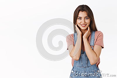 Attractive alluring brunette woman in dungarees, t-shirt touching her neck and smiling with gentle coquettish expression Stock Photo