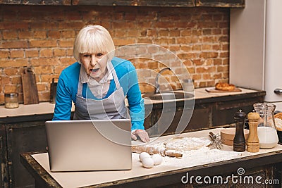 Attractive amazed shocked senior aged woman is cooking on kitchen. Grandmother making tasty baking. Using laptop Stock Photo
