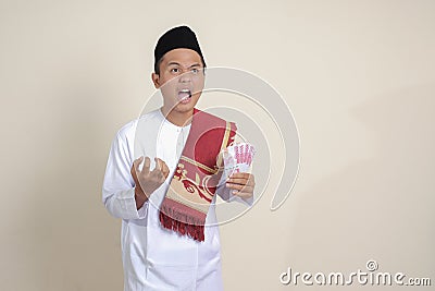 attractive Asian muslim man in white shirt making angry gesture showing one hundred thousand rupiah. Financial and Stock Photo