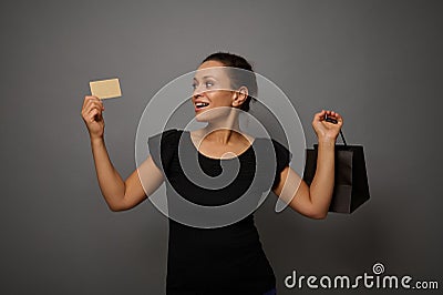 Attractive astonished mixed race beautiful woman poses against gray wall background with a shopping packet and rejoices looking at Stock Photo