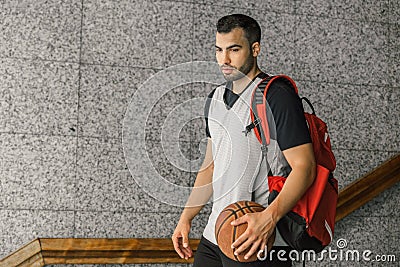 Attractive basketball player carries an orange ball and a red backpack, leaving the building where he lives Stock Photo