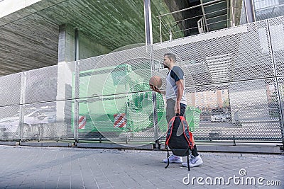 Attractive basketball player walks through an urban area carrying his red backpack and orange ball Stock Photo