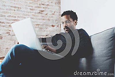 Attractive bearded African man working on laptop while sitting sofa at his modern home office.Concept of young people Stock Photo