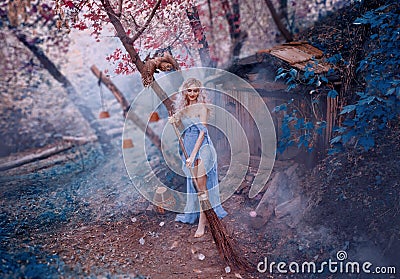 Attractive blond lady in long light dress of thin fabric with shoulder and open legs sweeps leaves with broom near Stock Photo