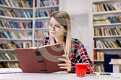 Attractive blond smart young student girl in eyeglasses sitting at the table, concentrated while reading book in the Stock Photo