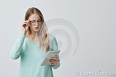 Attractive blonde female manager in stylsh eyewear works, look nervously at screen of tablet finds out unpleasant news Stock Photo