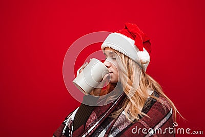 Attractive blonde girl in Christmas hat sings with closed eyes hot drink from big cup on red background, wears warm plaid. Winter Stock Photo