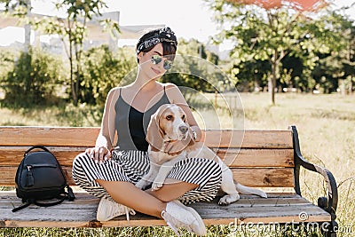 Attractive brunette lady in striped pants sitting with legs crossed and stroking beagle dog. Stylish smiling girl Stock Photo