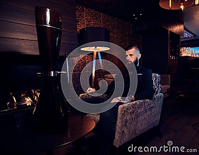 Attractive brutal man is sitting on the armchair and starting smoke hookah Stock Photo