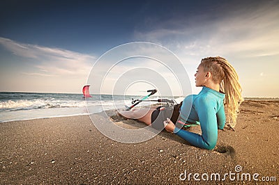 Attractive caucasian woman with dreadlocks on her head in a wetsuit lies on a sandy beach and holds her kite. Water Stock Photo