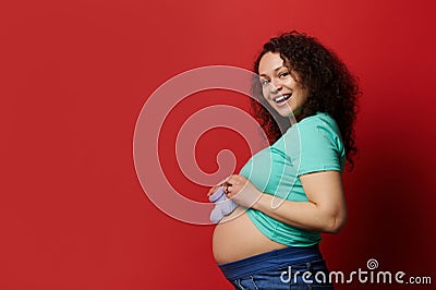 Attractive smiling ethnic pregnant woman with bare belly, posing with cute blue knitted baby booties, red background. Stock Photo