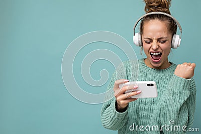 Attractive emotional positive young woman wearing blue sweater isolated over blue background wearing white bluetooth Stock Photo