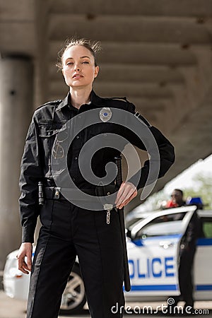 attractive female police officer in uniform looking at camera with blurred partner near car Stock Photo