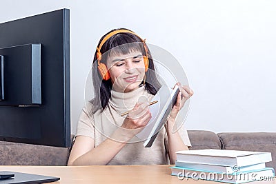 An attractive female teacher in orange headphones sitting in front of a computer monitor, explains an online task Stock Photo