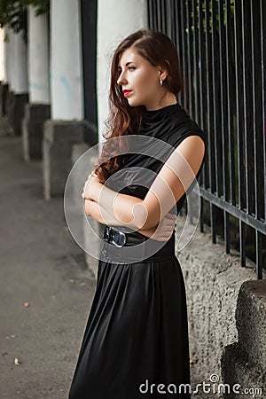 Attractive girl in a black dress with a corset on a background of a fence with clones. Evening walk on the city street. Stock Photo