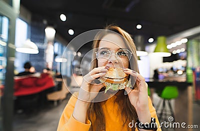 Attractive girl in orange clothes and glasses sits in a fast-food restaurant and eats a great appetizing burger Stock Photo