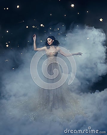 An attractive goddess stands in the clouds in a luxurious, gold, sparkling dress. Whimsical hairstyle. Against the Stock Photo