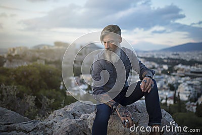 Attractive hipster with a grey beard is sitting on a rock high over Athens City with Acropolis view Stock Photo