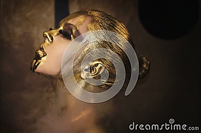 Attractive hot woman with beautiful liquid gold on her face and body is posing dark background in smoke, closed eyes Stock Photo