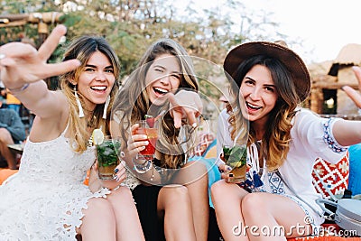 Attractive long-haired girls in stylish attires and accessories drink cocktails and posing with hands waving. Portrait Stock Photo