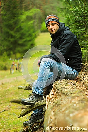 Attractive male cyclist with unshaven in a sports jacket, blue jeans, a buff and trekking boots sits on a tree trunk in the autumn Stock Photo