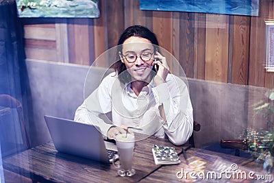 Attractive man in white shirt talking by phone and working on laptop in cafe or restaurant, having rest drinking coffee latte Stock Photo