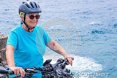 Attractive mature cyclist woman enjoying activity at sea with her electro bike. Standing on the cliff Stock Photo