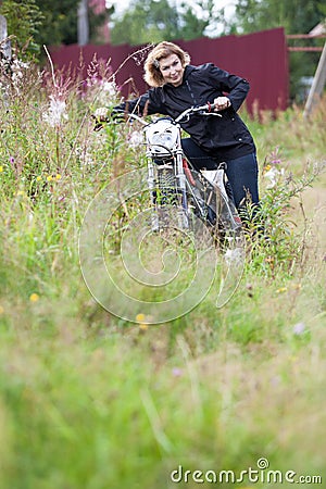 Attractive mature woman posing with motocross motorcycle in high grass, holding the wheel and looking at camera Stock Photo