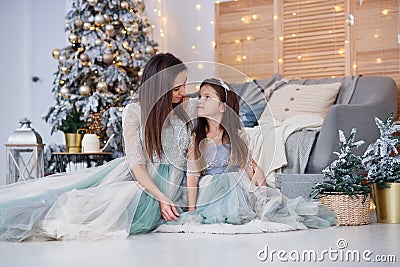 Attractive mom and her cute daughter in a gentle evening dresses sitting on the floor near the Christmas tree in a cozy Stock Photo