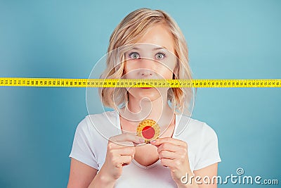 Attractive sad blonde woman with make-up and lips color fuchsia hold a high-calorie cookie biscuit iwith measuring tape Stock Photo