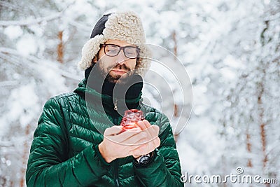 Attractive serious male wears warm winter clothes, keeps candle, stands against winter forest background, enjoys fresh air, being Stock Photo