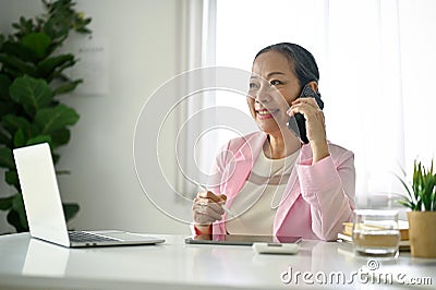 Attractive senior Asian businesswoman sits at her desk and talks on the phone Stock Photo