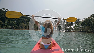 Attractive sportive blonde woman with sunglasses rows pink plastic kayak putting up paddle on sea against hills with Stock Photo