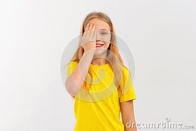 Attractive teen girl with blonde hair, covering half of her face with hand, looks with one eye at camera with happy face, stands Stock Photo