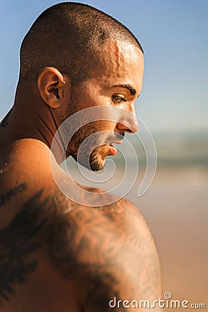 An attractive topless man with tattoos standing on a beach next to a sea Stock Photo