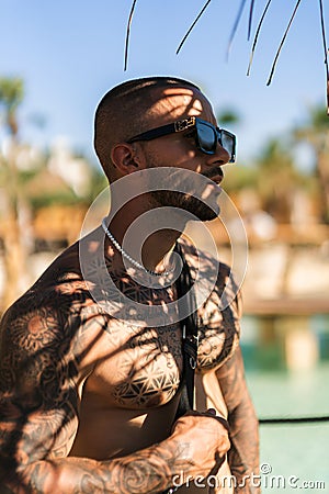 An attractive topless man with tattoos standing next to a sea under a tree Stock Photo