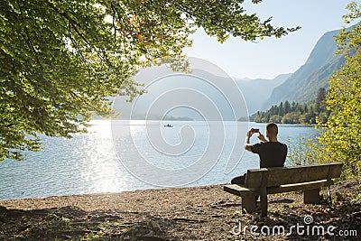 Attractive tourist with phone camera taking picture of beautiful lake, male enjoying vacation journey in summer Stock Photo