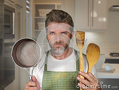 Attractive unhappy and overwhelmed home cook man in apron holding spoon and pan feeling upset and lazy at house kitchen in Stock Photo