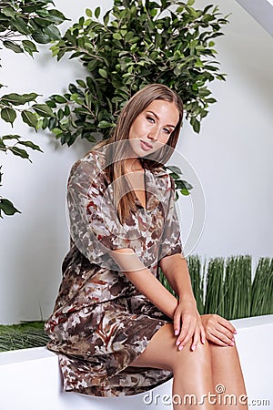 An attractive woman in a khaki dress with camouflage print sits in the office with green plants. Stock Photo