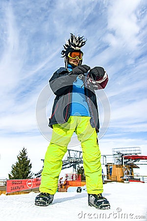 Attractive youg man tourist in funny mohawk hat, green pants, ski glasses mask and black mittens on a background of blue Stock Photo