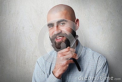 Attractive yound balded barber with thick black beard and mustache smiling while holding straight razor on his beard. A young hair Stock Photo
