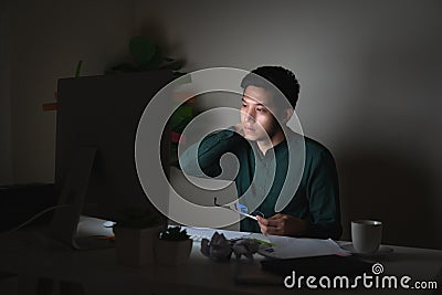 Attractive young asian man working late dark night looking at laptop computer in dark home office desk feeling tired on work load Stock Photo