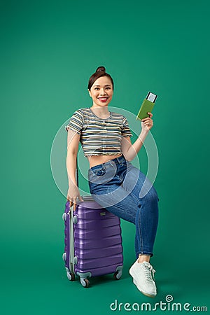 Attractive young Asian woman sitting on suitcase and holding passport, ticket flight over green background Stock Photo