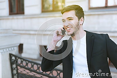 Attractive young business man using mobile phone on building outdoors. Cheerful handsome caucasian man in black suit in the city. Stock Photo