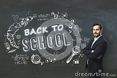 Attractive young businessman with folded arms standing on creative back to school sketch on blackboard background. Education, Stock Photo