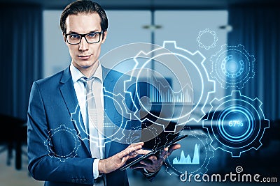 Attractive young european businessman with digital pad and glowing cogwheel and chart hologram on blurry office interior Stock Photo