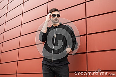 Attractive young hipster man in fashionable black clothes straightens trendy sunglasses. Stylish urban model guy stands outdoors Stock Photo