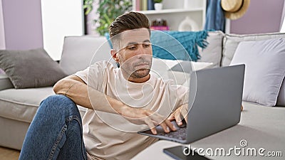 Attractive young hispanic man, concentrated and serious, sitting on the floor in his cozy apartment, working online on his laptop, Stock Photo